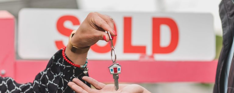 House key with a home-shaped keychain handed over in front of a red SOLD sign, illustrating Aldar villa sales in Abu Dhabi