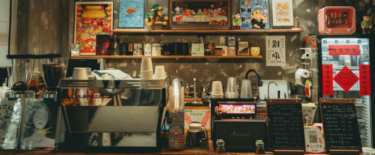 Coffee shop counter in China with an espresso machine, stacks of takeaway cups, handwritten menu boards and a QR code: Mubadala is investing about USD 1 billion in Luckin Coffee