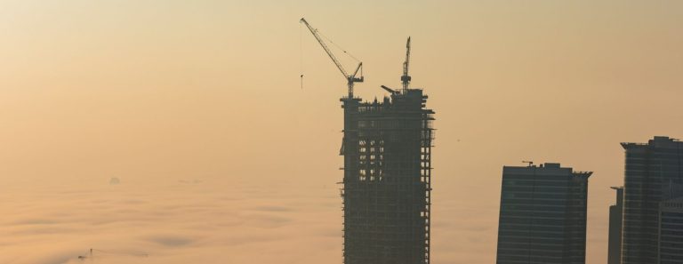 High-rise tower under construction with tower cranes on top, rising above morning fog next to glass office towers in Dubai