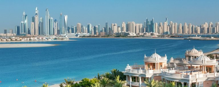 Dubai Marina skyline seen from Palm Jumeirah, with frond villas across the water and a palatial beachfront building in the foreground