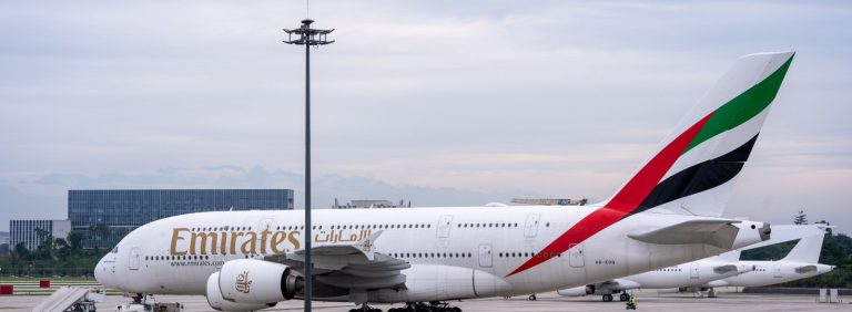 Emirates Airbus A380 parked on the apron with the terminal and other aircraft behind it, illustrating the 1.5 billion passengers handled by Dubai International since 1960 and the planned move of operations to Al Maktoum International