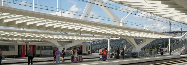 Passengers waiting on a sunlit platform of a modern railway station with a wide white canopy and intercity carriages, illustrating the launch of the Etihad Rail passenger service between Dubai and Abu Dhabi