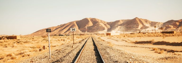 Single track railway line running through desert scrub towards bare rocky mountains, illustrating the 238 km Hafeet Rail link between the UAE and Oman
