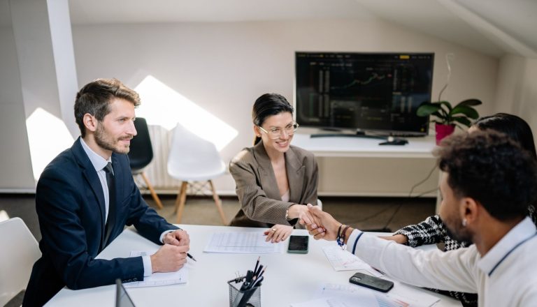 Business professionals shaking hands across a meeting table with financial charts and a market screen