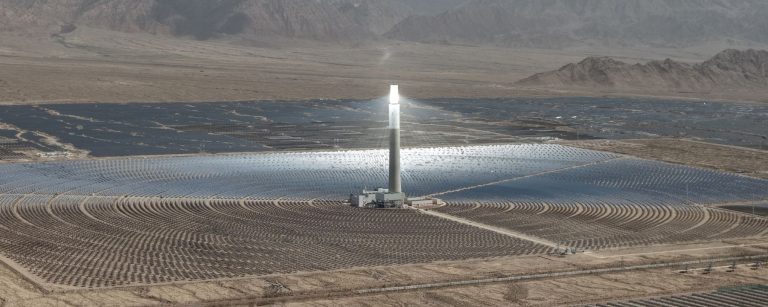 Concentrated solar power tower with a glowing receiver surrounded by a circular field of heliostat mirrors and photovoltaic panels in the desert