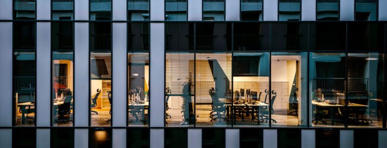 Illuminated open-plan office floor behind a glass facade at dusk, illustrating the State Street operating centre planned in Al Ain by the end of 2027