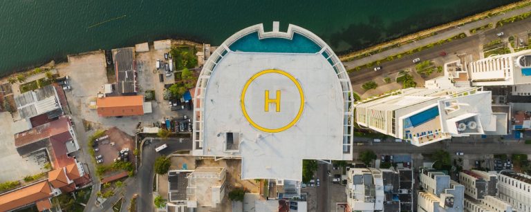 Aerial top-down view of a rooftop helipad with a yellow H marking on a seafront high-rise building