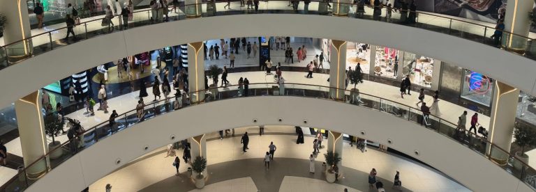 Shoppers walking across several levels of a large multi-storey shopping mall atrium in Dubai, illustrating the forecast growth of UAE retail sales to USD 129.5 billion by 2030