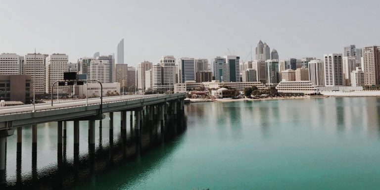 Road bridge on piers over turquoise water leading to the Abu Dhabi skyline, illustrating island road links such as the Mid-Island Parkway