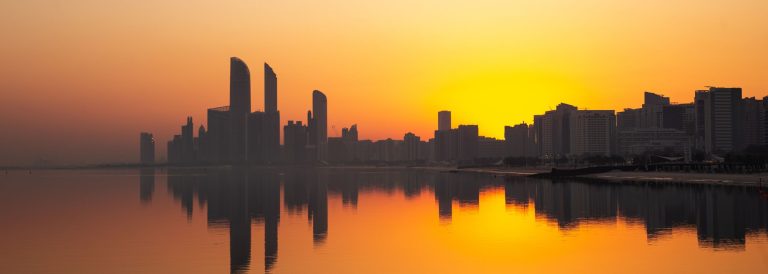 Abu Dhabi skyline silhouetted against an orange sunset and reflected in calm water