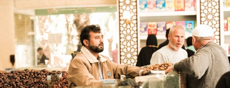Traders negotiate over loose dates at a food market stall, a seller in front of open crates of dates