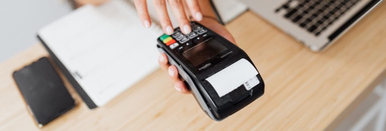 A merchant holding a card payment terminal on a desk next to a laptop and a smartphone, in-store and online payment acceptance