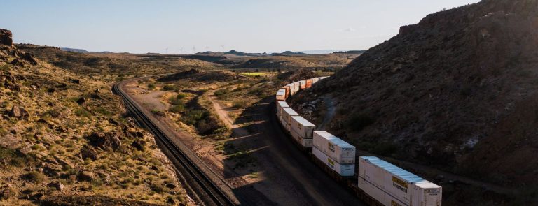 Double-stacked container freight train on a curving railway through a desert canyon with wind turbines on the horizon