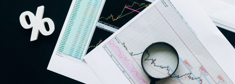 Percent sign, printed financial charts and a magnifying glass on a dark desk